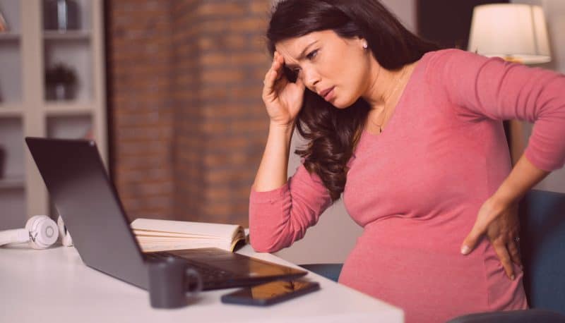 Pregnant woman stressed with headache wearing a pink top working from home on laptop for workplace discrimination