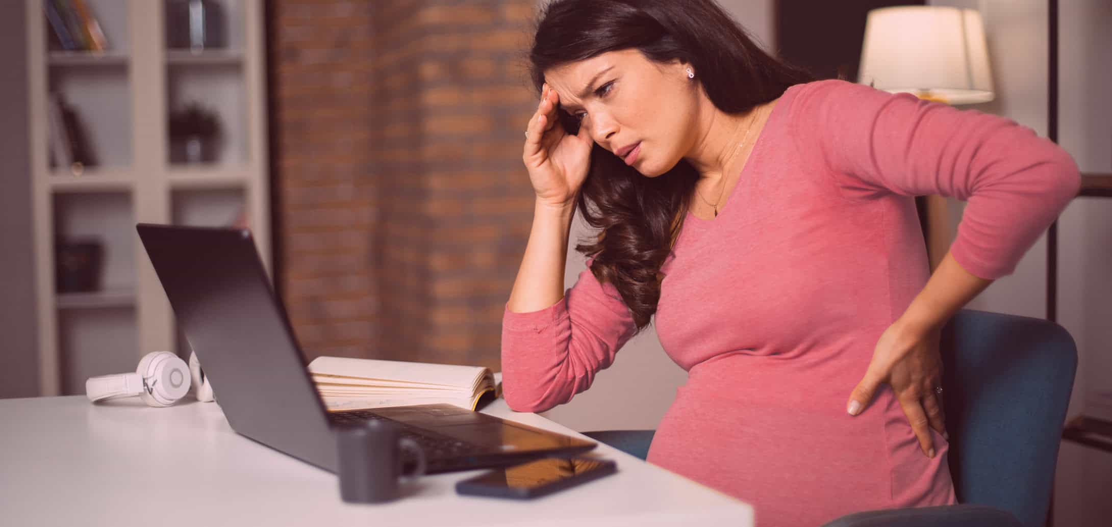 Pregnant woman stressed with headache wearing a pink top working from home on laptop for workplace discrimination