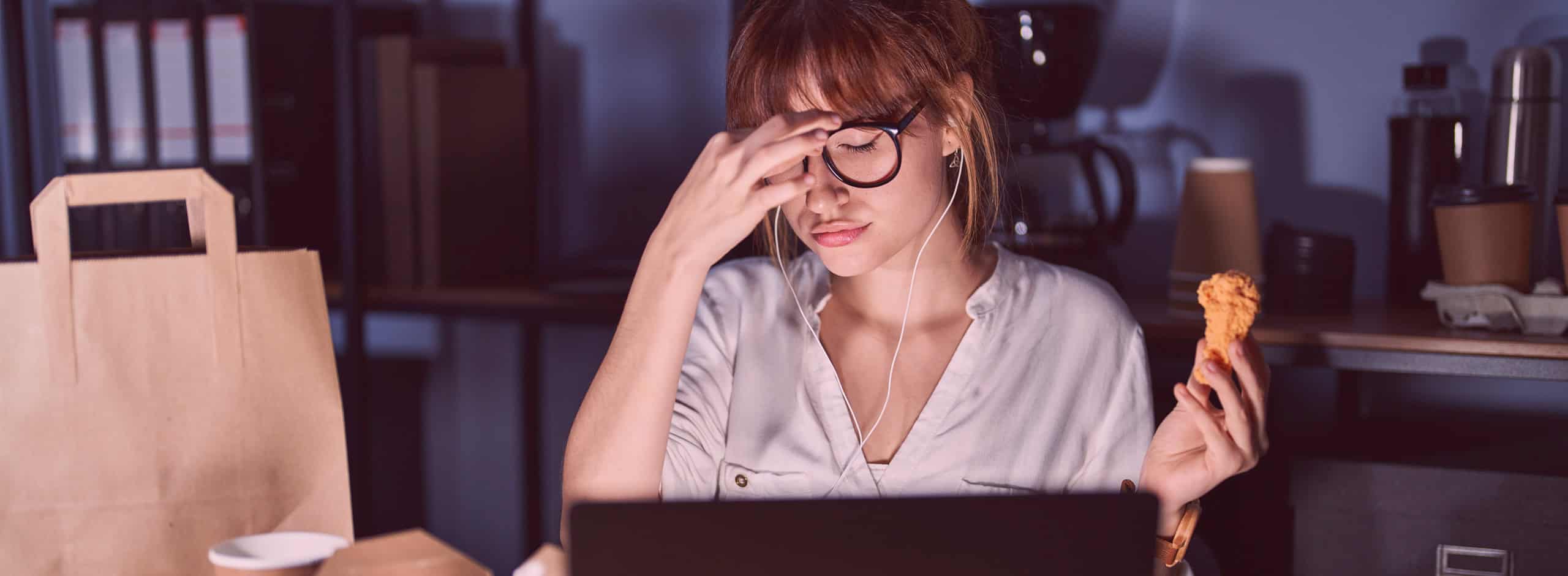 woman working late sitting at laptop with dinner holding chicken leg and pinching nose wearing glasses, room is dark