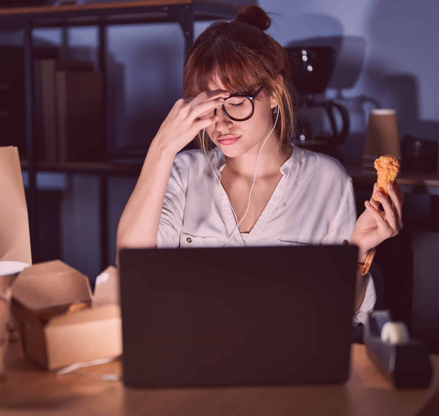 Woman working late sitting at laptop with dinner holding chicken leg and pinching nose wearing glasses, room is dark