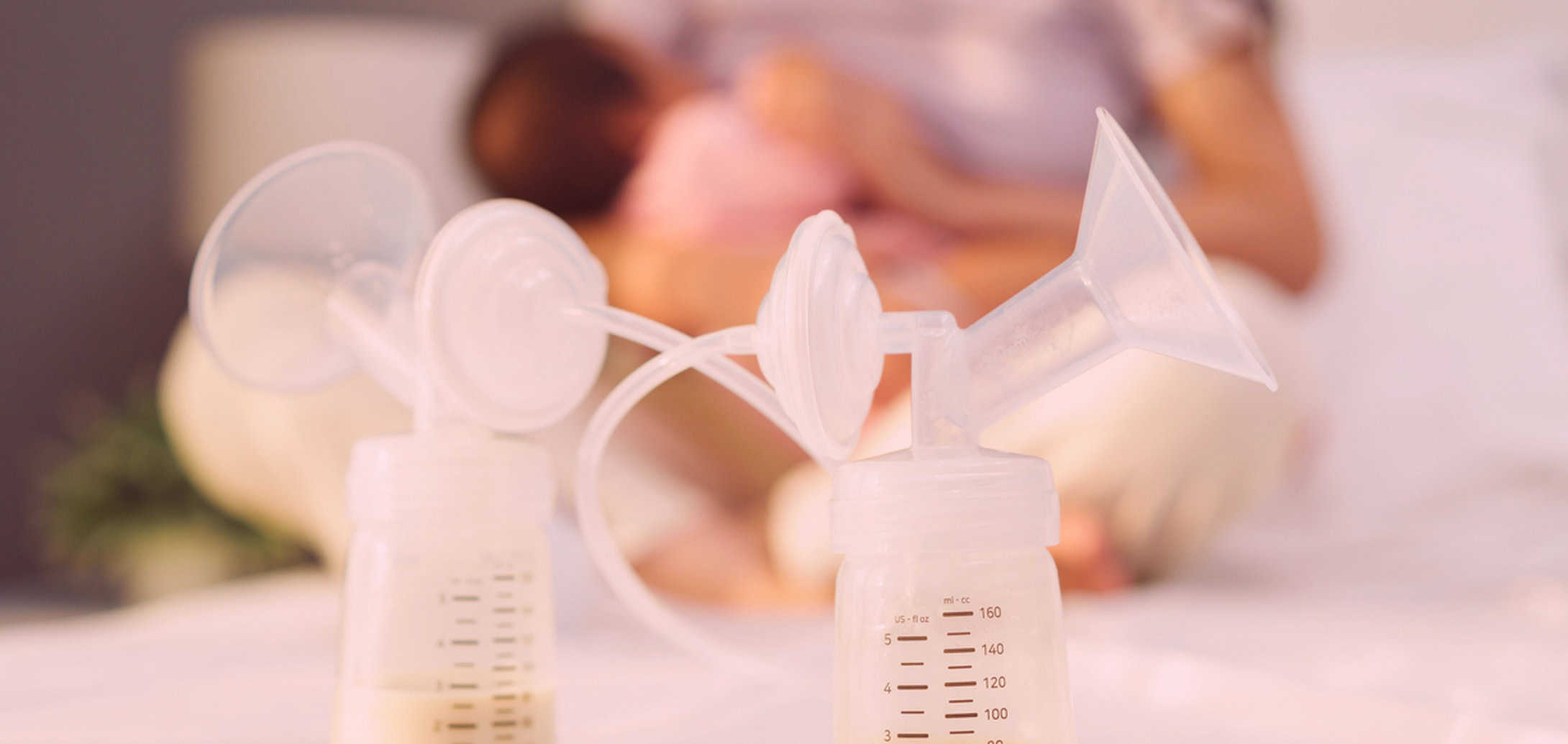 Two breast pumps in foreground, nursing mother sitting out of focus in background