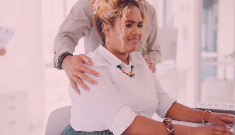 Woman sitting at her desk working on a computer, looking very uncomfortable as a coworker touches her shoulders.