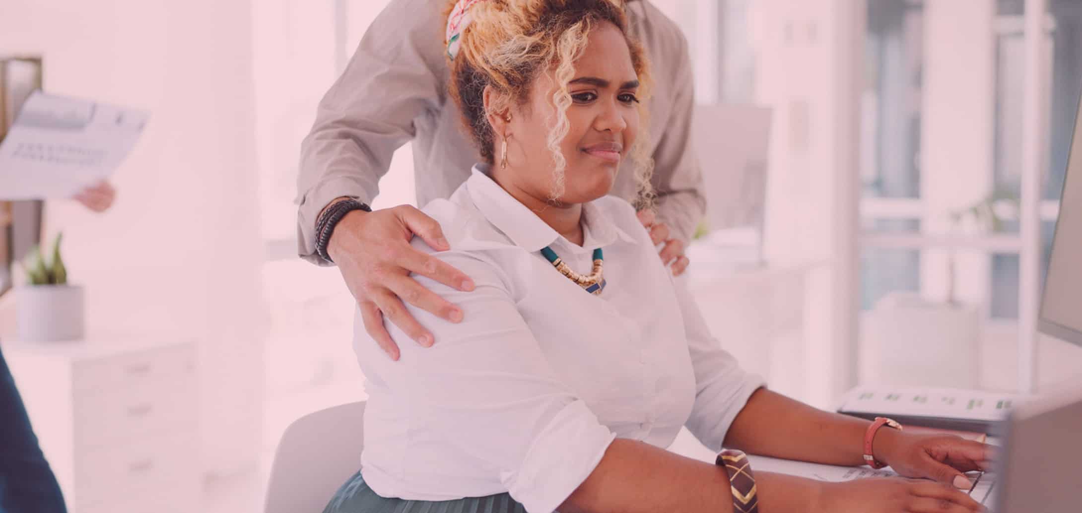 Woman sitting at her desk working on a computer, looking very uncomfortable as a coworker touches her shoulders.