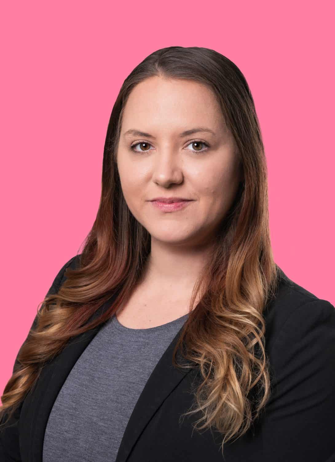 Alicia Brush attorney wearing a black blazer and gray tee shirt with long hair on pink background