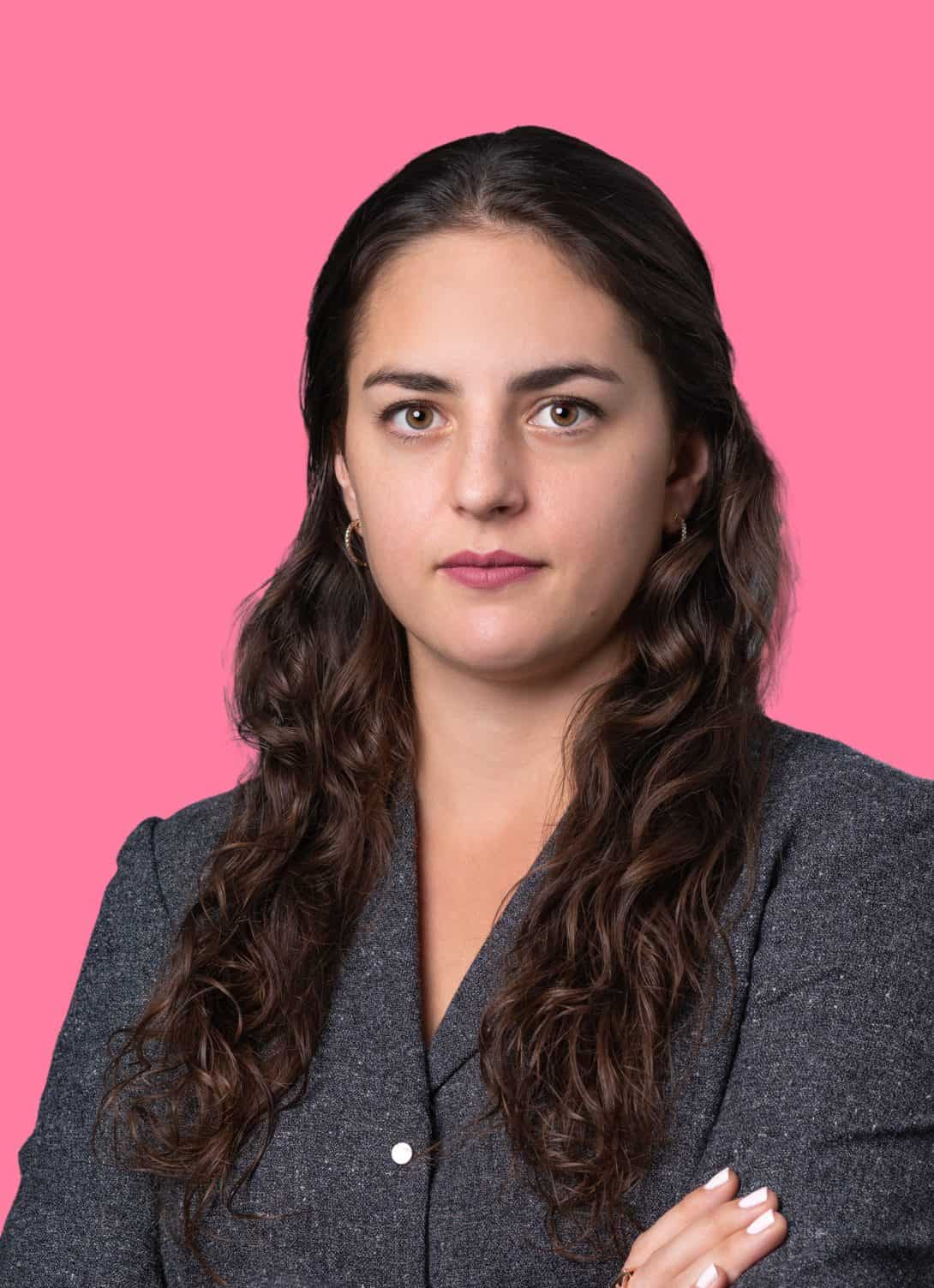 Hagit Goltzer attorney at lady justice chest up portrait wearing gray blazer, looking at camera, on pink background, long wavy brunette hair