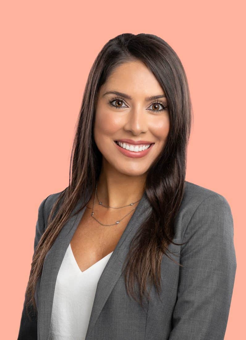 Tara Zabehi portrait from chest up wearing gray blazer and white top, long straight hair, smiling at camera on peach background