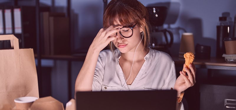 Woman working late sitting at laptop with dinner holding chicken leg and pinching nose wearing glasses, room is dark