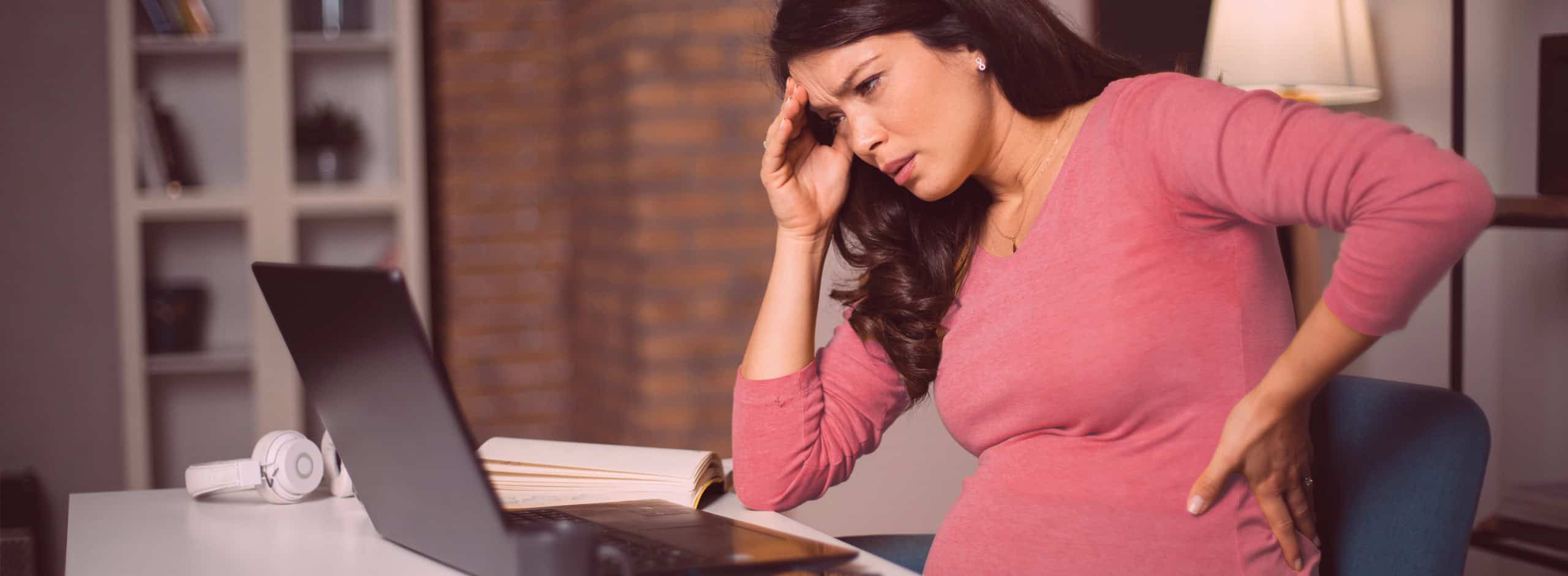 Pregnant woman stressed with headache wearing a pink top working from home on laptop for workplace discrimination