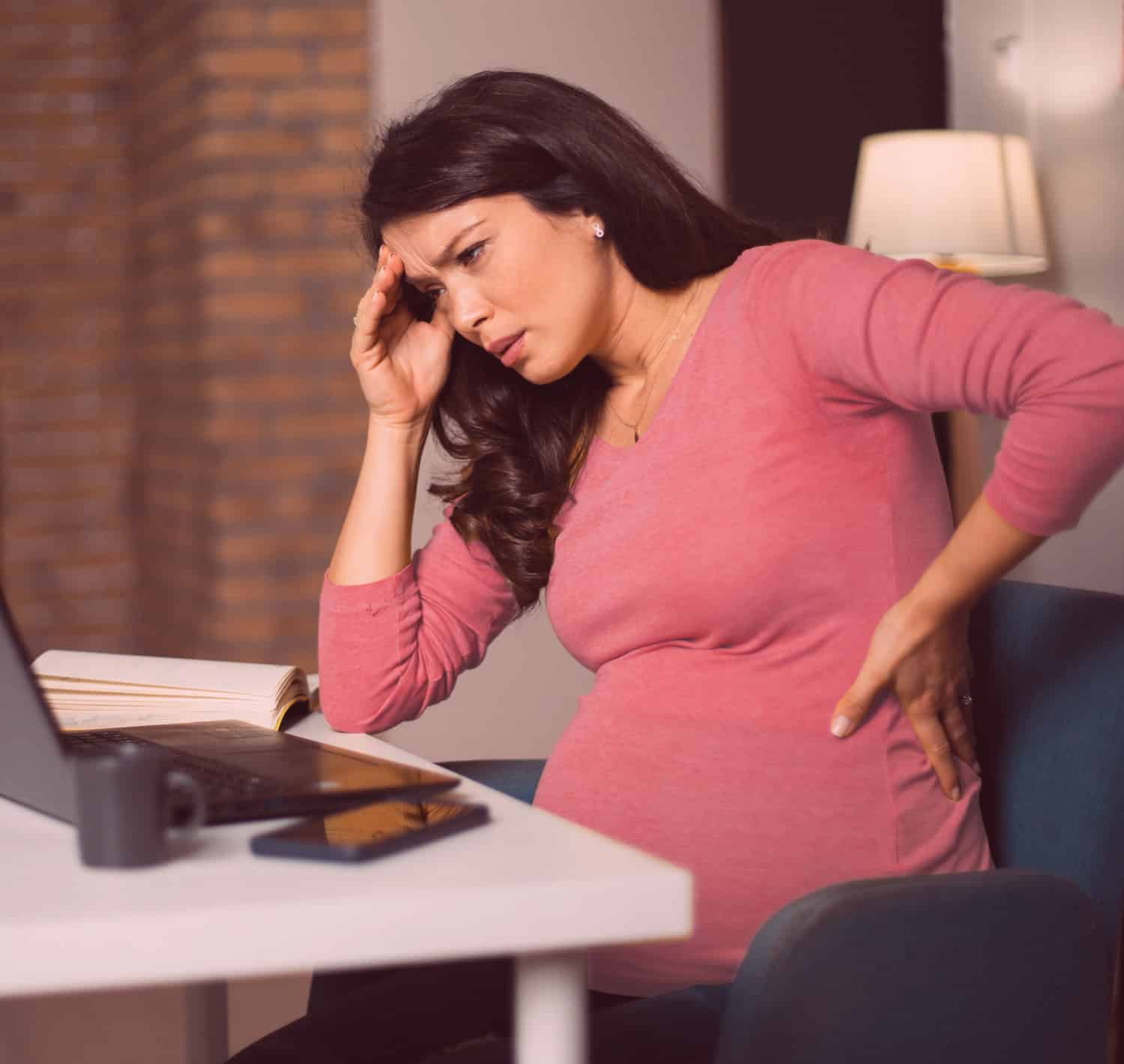 Pregnant woman stressed with headache wearing a pink top working from home on laptop for workplace discrimination