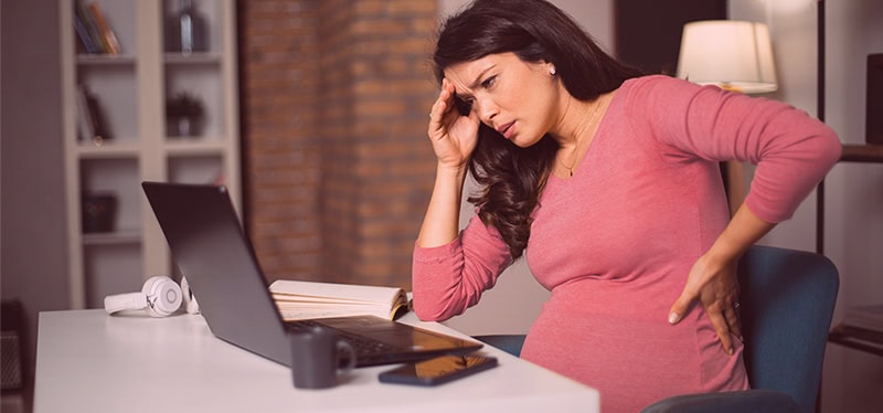 Pregnant woman stressed with headache wearing a pink top working from home on laptop for workplace discrimination